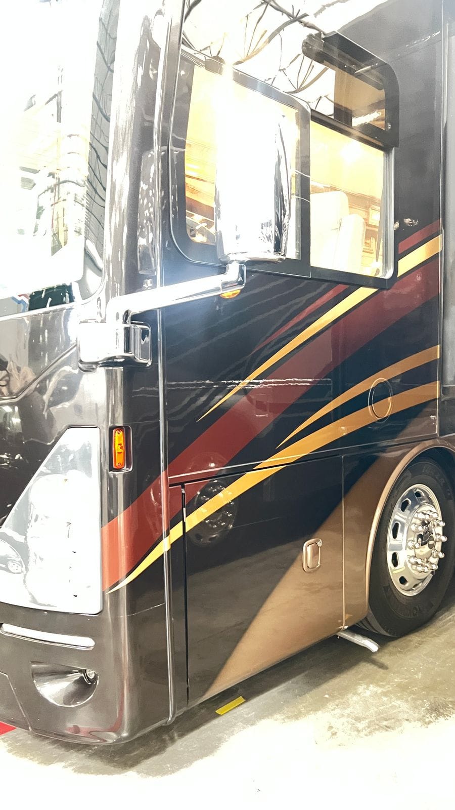 2016 Thor Tuscany 45AT driver-side view at the cab door with chrome west-coast mirror, sliding driver window, gold and burgundy side stripes over near-black paint and a polished aluminum wheel