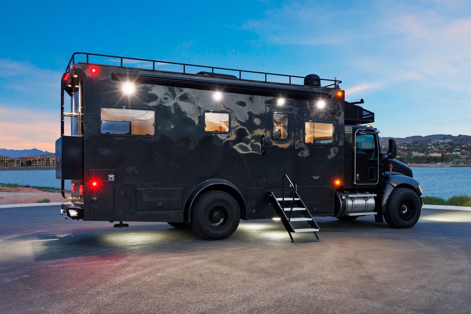 2017 Global Expedition Vehicles The Beast rear passenger-side three-quarter at dusk with the entry stairs down and exterior lighting lit, showing the length of the box body and the rear corner ladder