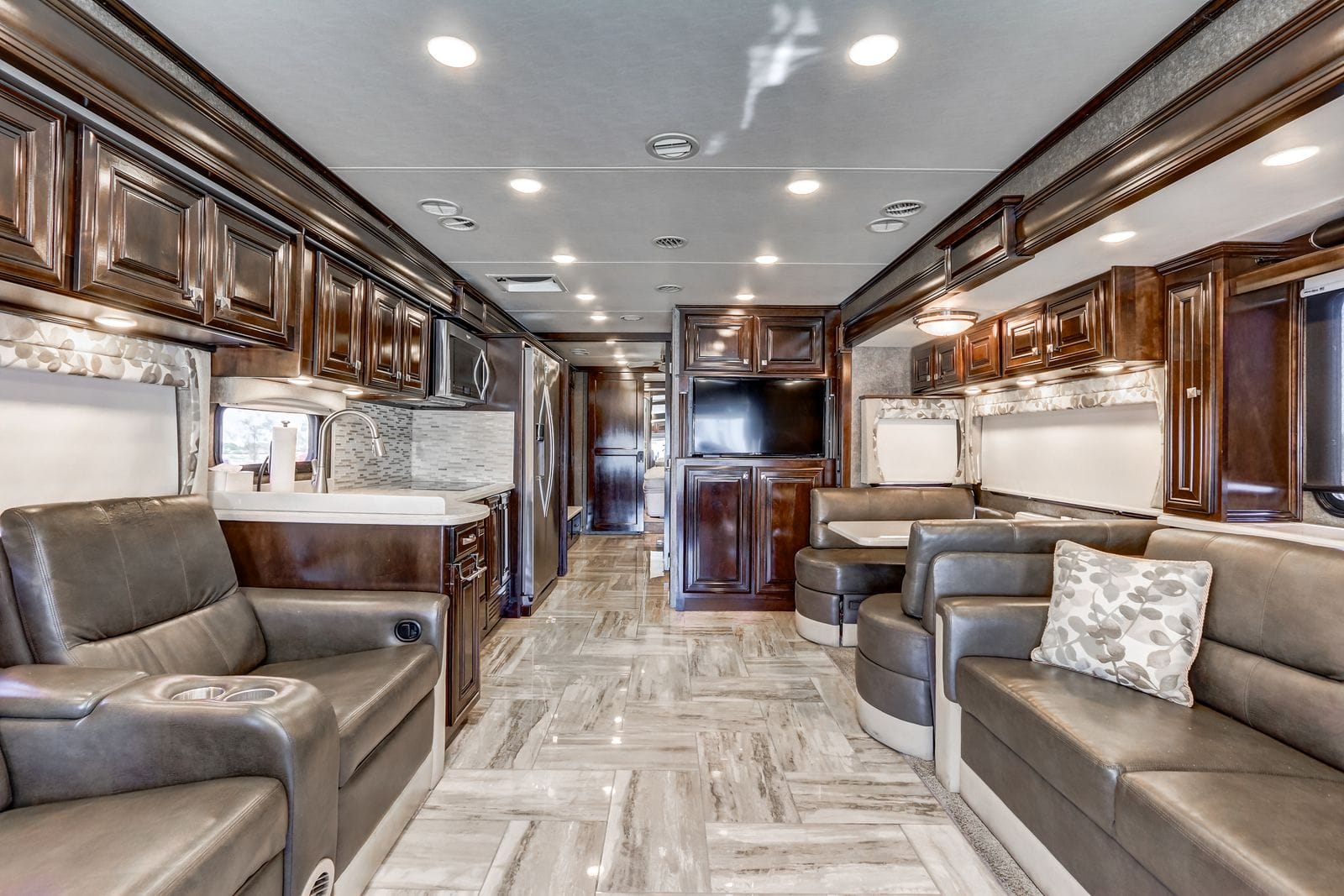 2017 Thor Venetian 40A interior looking aft down the centre aisle with high-gloss dark wood cabinetry, leather sofa, galley with stainless refrigerator and microwave, flat screen television and booth dinette