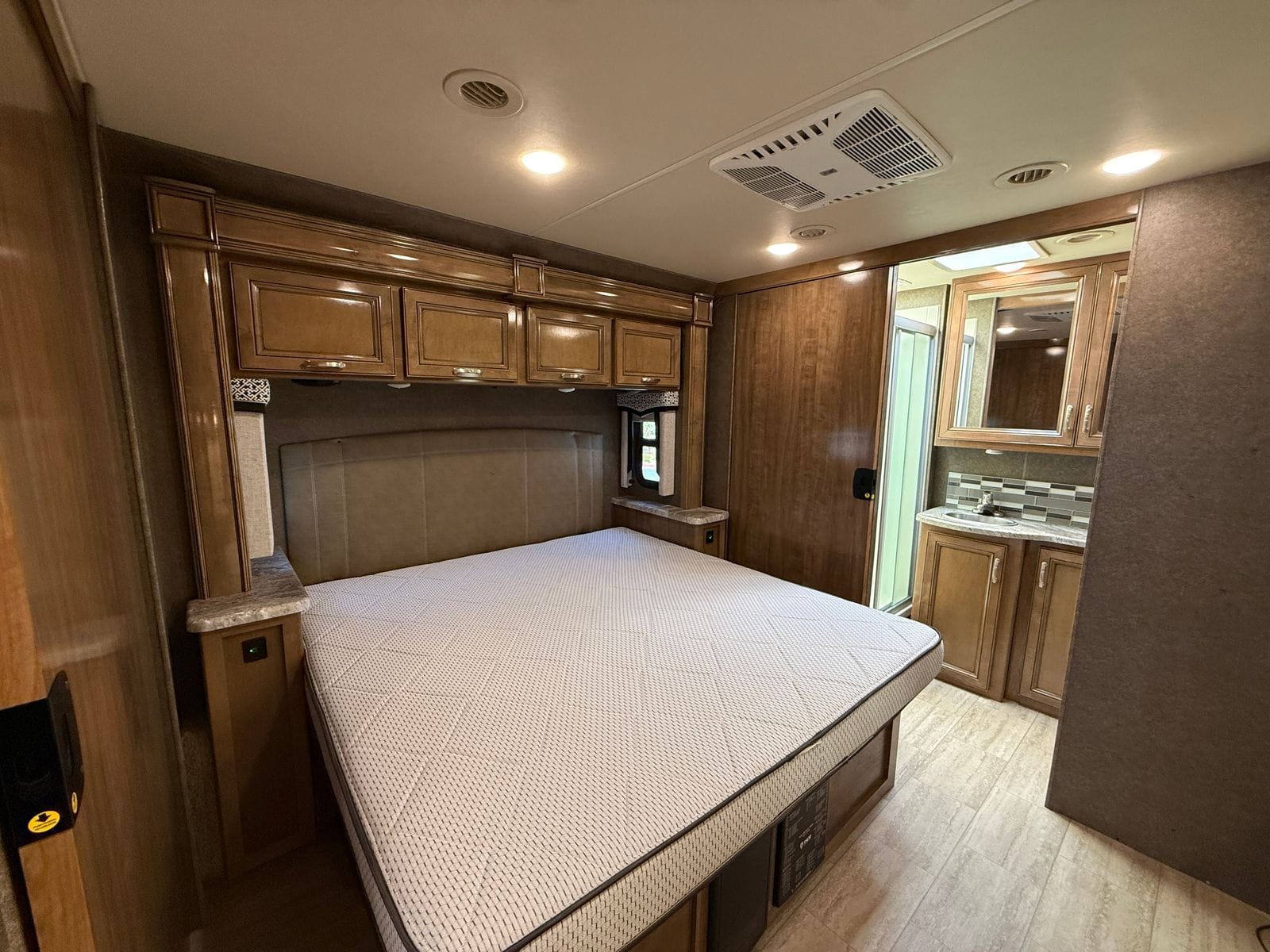2020 Thor Magnitude BH35 rear bedroom, showing the bed on its raised platform with a storage drawer beneath, overhead cabinets, nightstands either side and the rear bath through the doorway at right