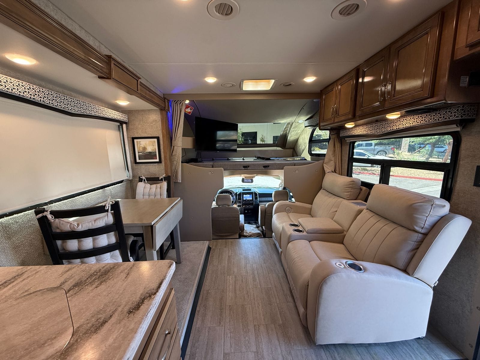 2020 Thor Magnitude BH35 living area looking forward, showing the theater seating on the right, the freestanding dinette on the slide-out at left, the galley counter in the foreground and the cab-over bunk above the cockpit