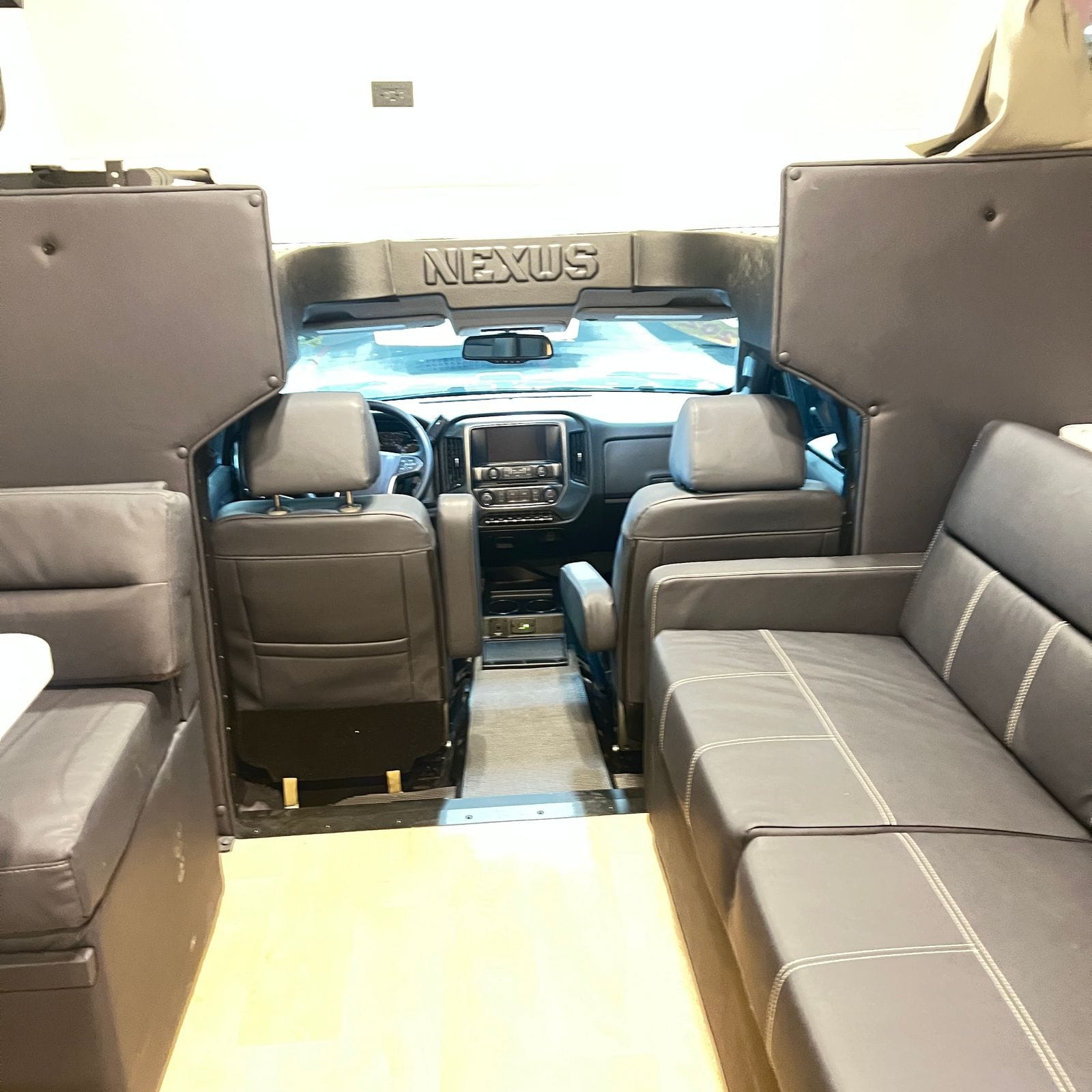 2026 Nexus Rebel 35R view forward from the living area through the cab, showing the embossed Nexus dash cap, two grey captain's chairs and flanking sofa and dinette seating