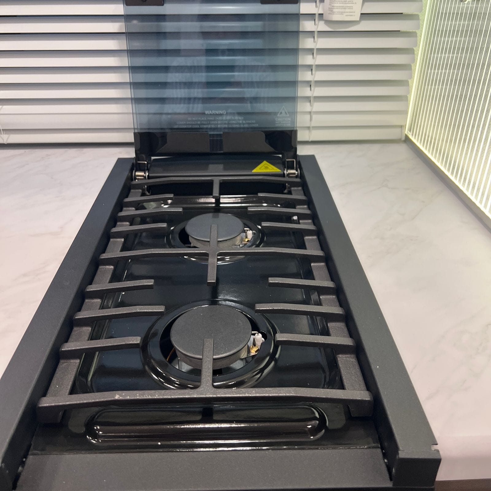 2026 Thor Delano 24XL overhead view of the two-burner gas cooktop with cast grates, black burner caps and the raised glass cover against the marble-look counter