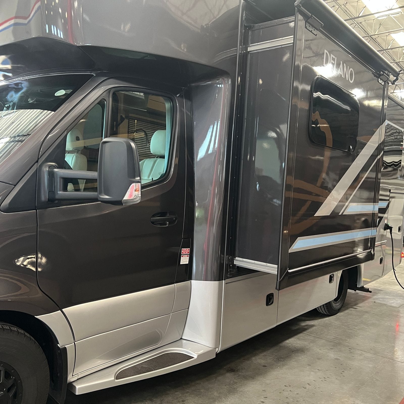 2026 Thor Delano 24XL driver-side view from the front corner showing the cab door and heated remote mirror, the extended slide-out room with awning rail and Delano script on the coach side