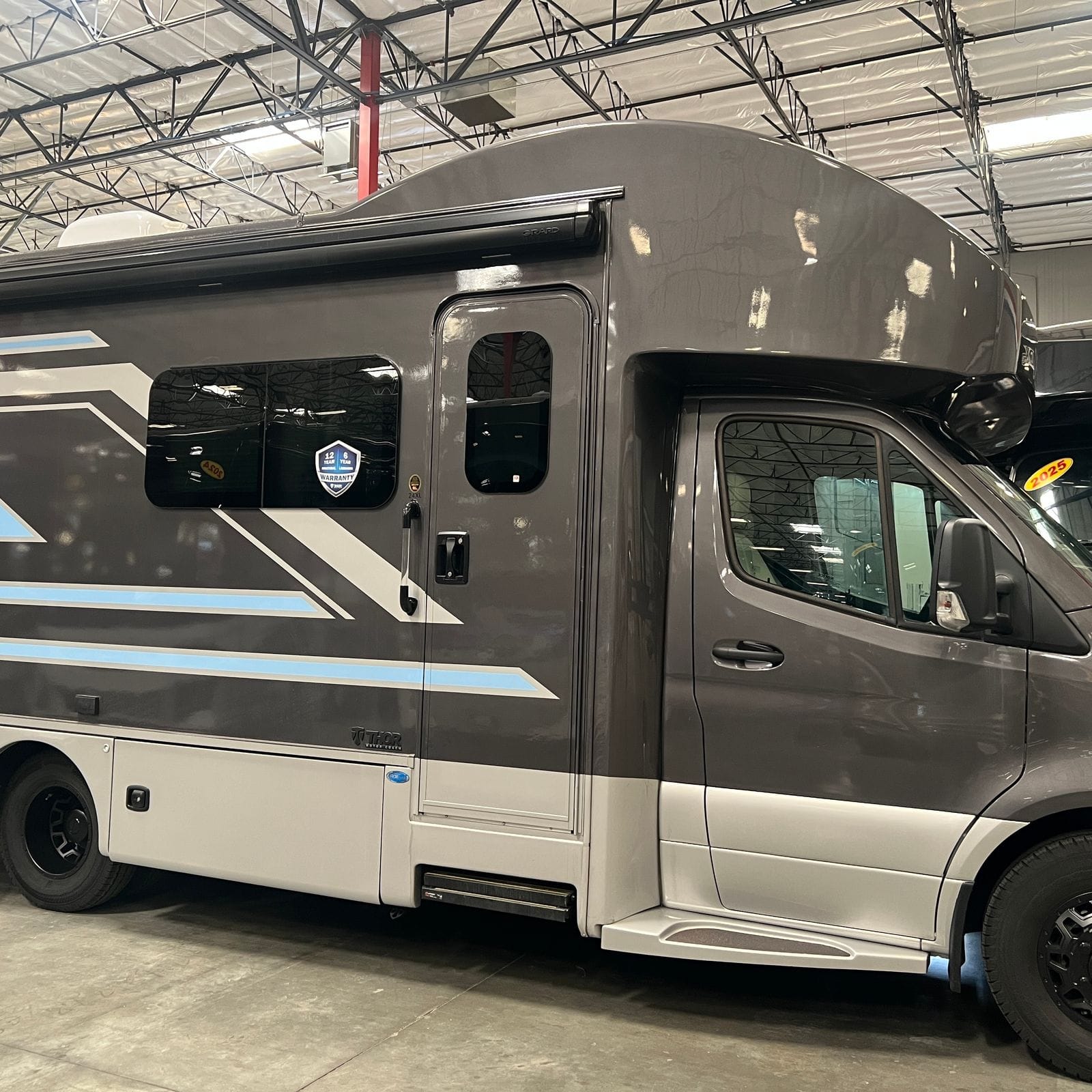 2026 Thor Delano 24XL passenger-side view showing the entry door with grab handle, tinted frameless windows, grey and light-blue swoosh graphics over a silver lower skirt and black wheels