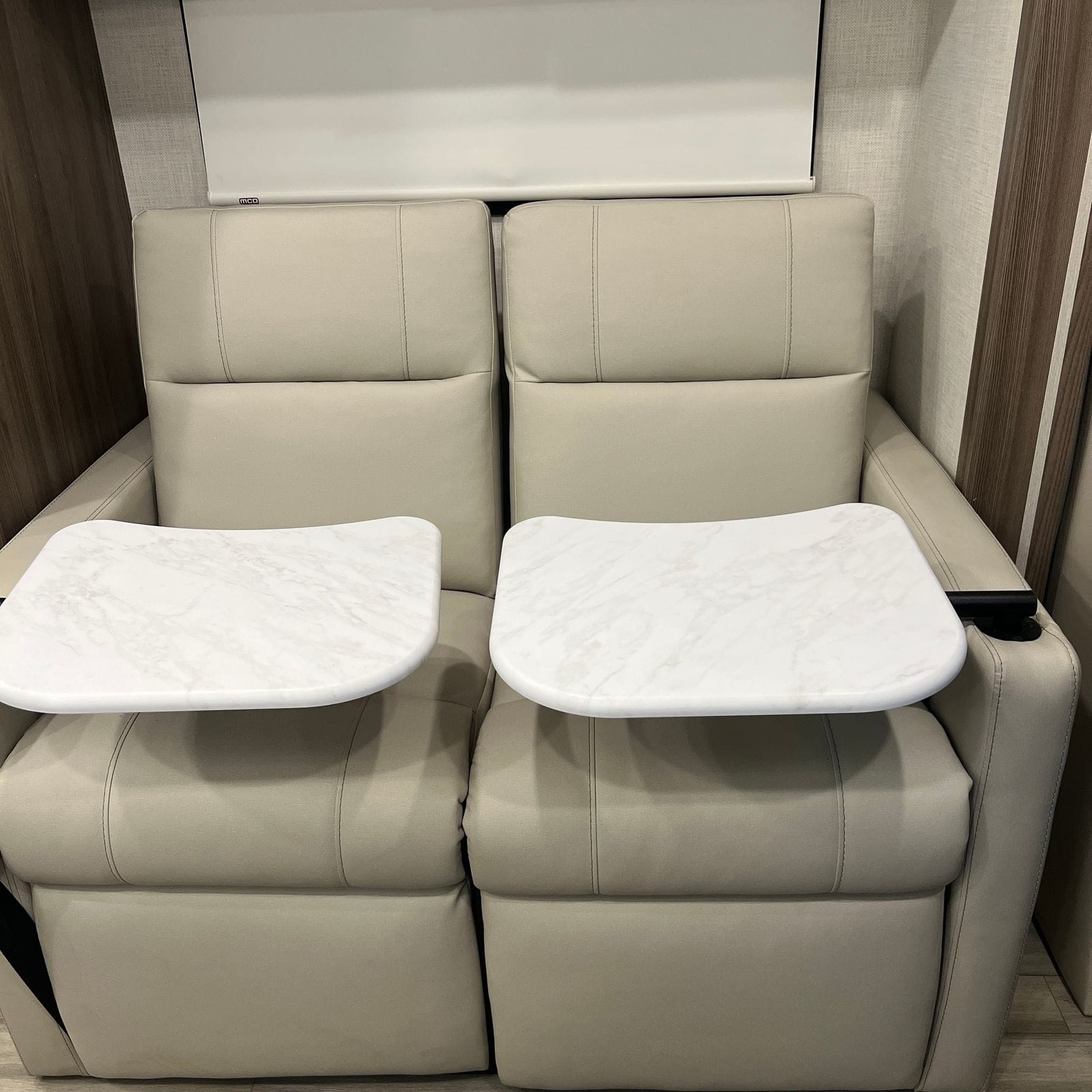 2026 Thor Delano 24XL two tan leatherette theater seats with deployed marble-look swing-out side tables, a roller shade behind and wood-grain wall panels either side