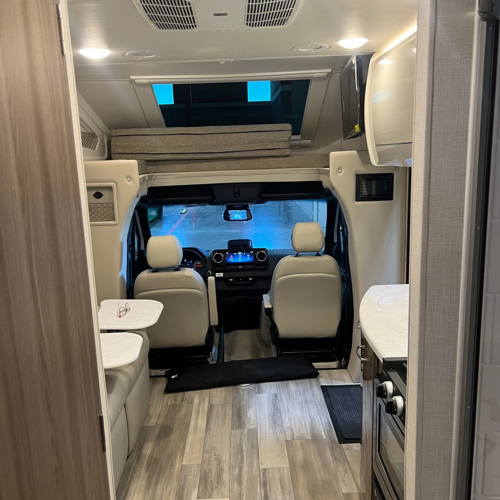 2026 Thor Delano 24XL looking forward down the aisle from the rear showing wood-plank vinyl flooring, theater seats at left, galley counter at right and the cab-over bunk with skylight
