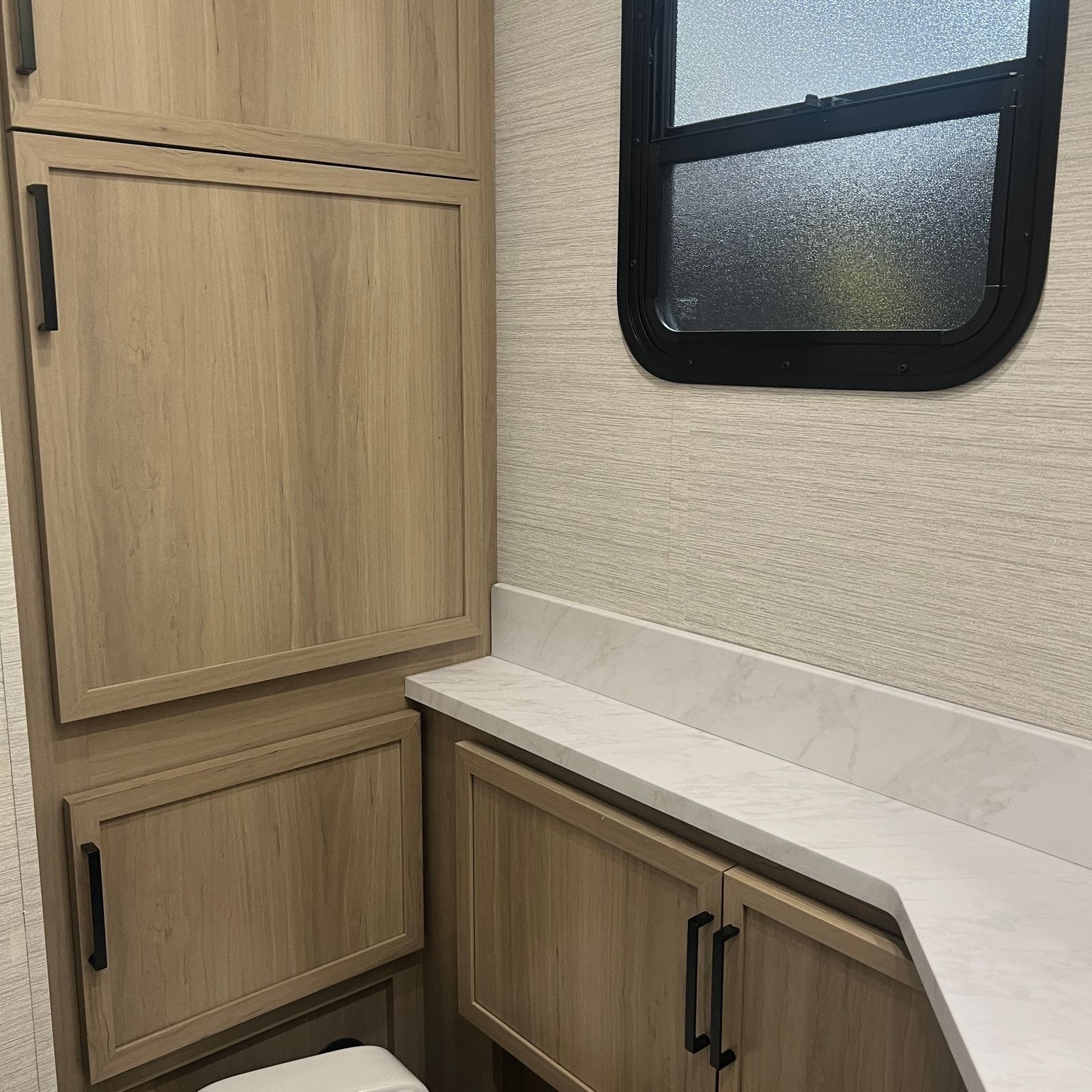 2027 Dynamax Europa S 37CA bathroom showing light wood linen cabinets, a frosted privacy window and an L-shaped marble-look counter with backsplash