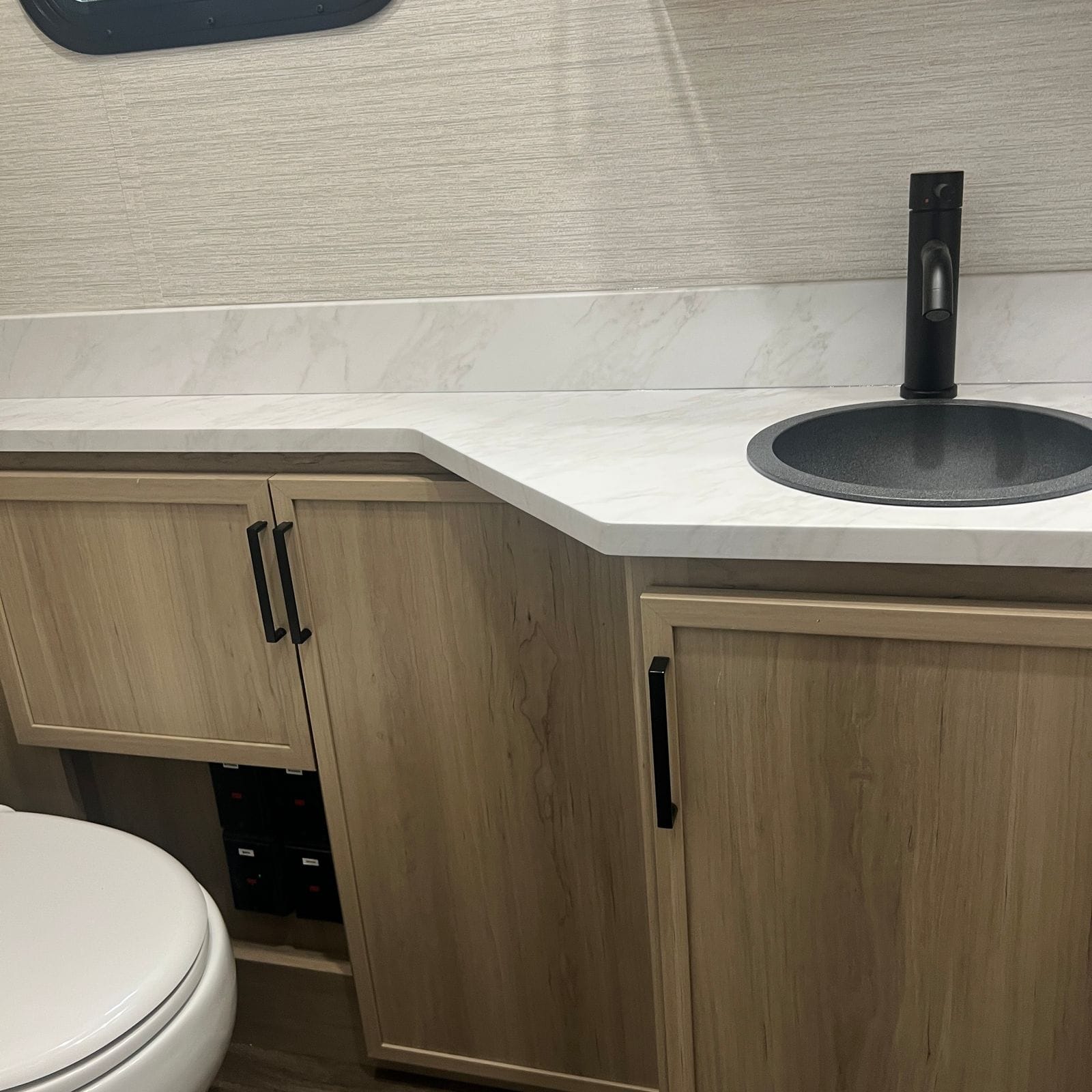 2027 Dynamax Europa S 37CA bathroom counter run with the round grey sink and black faucet, light wood cabinet doors with black pulls and the toilet alongside