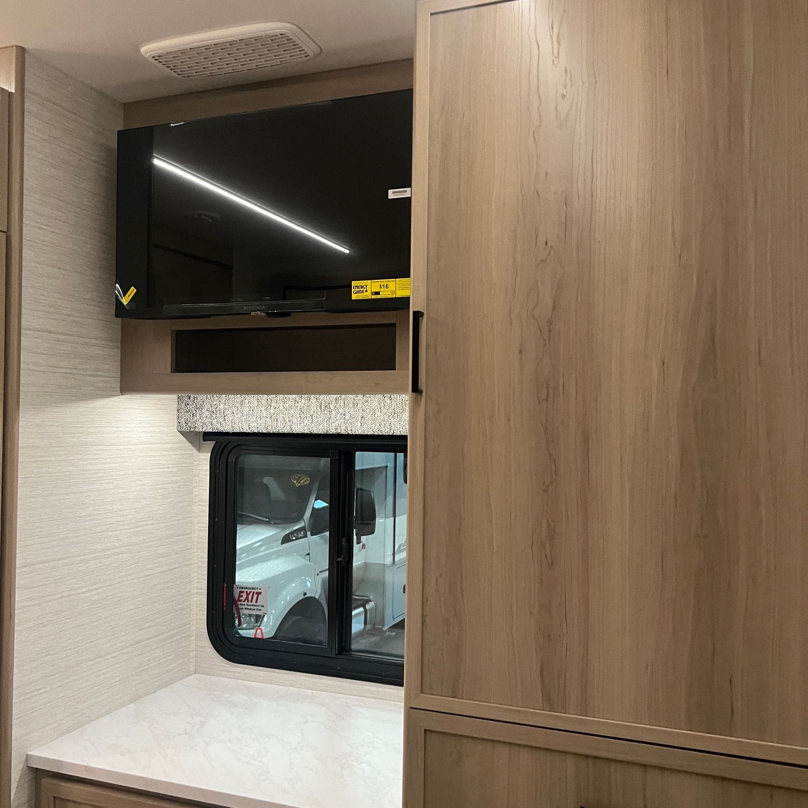 2027 Dynamax Europa S 37CA bedroom wall with a wall-mounted flat-screen television, a storage cubby, valanced side window with emergency exit label and a marble-look nightstand surface below