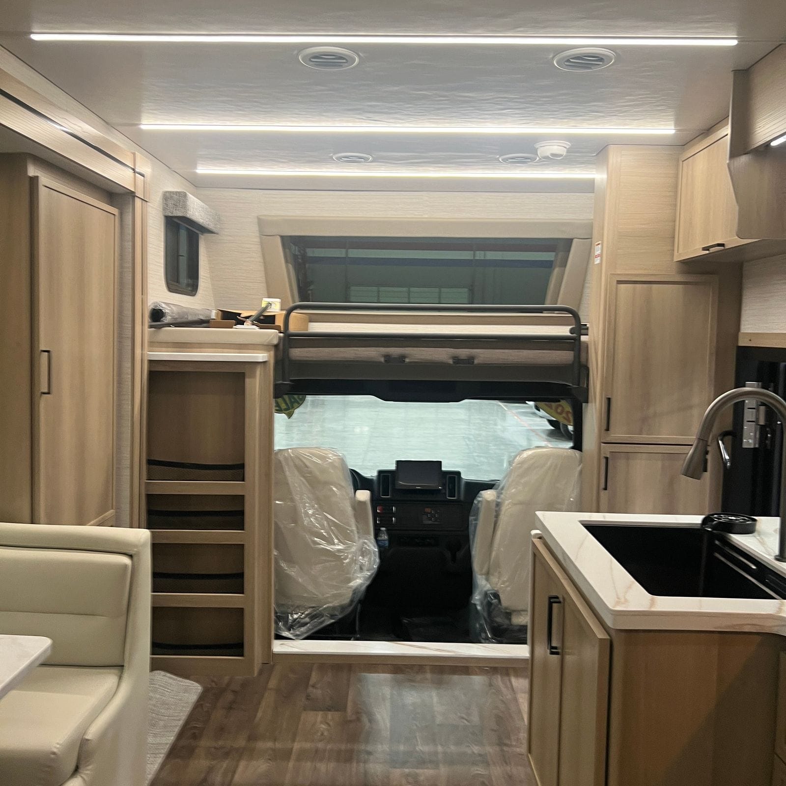 2027 Dynamax Europa S 37CA interior looking forward from the living area toward the cockpit, showing the overhead bunk with its safety rail, the galley sink at right and the dinette at left