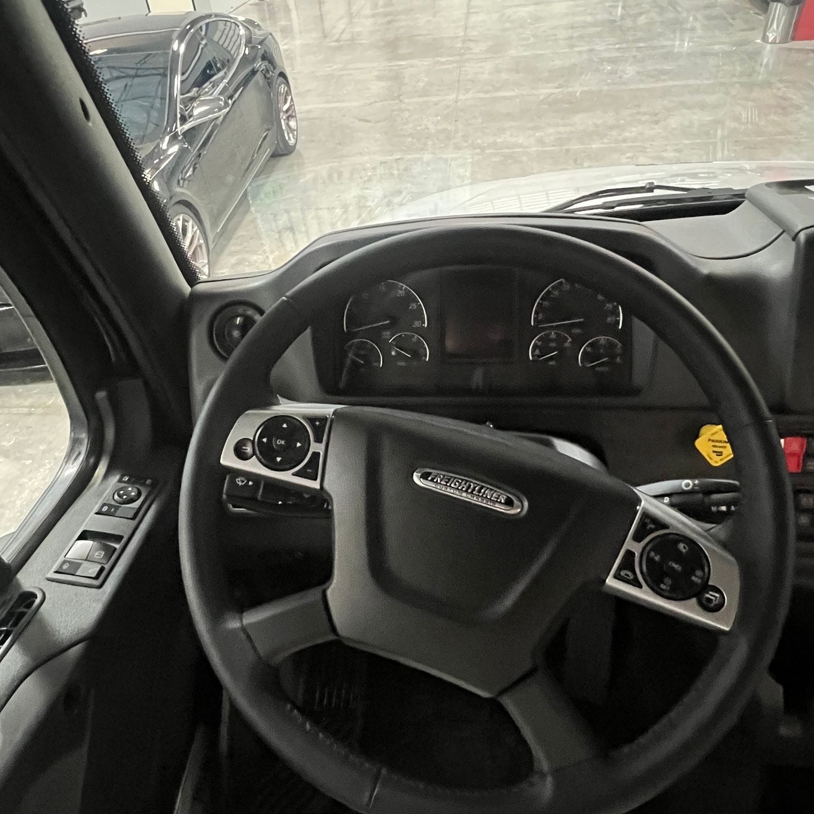 2027 Dynamax Europa S 37CA driver position showing the Freightliner steering wheel with multifunction controls, analog gauge cluster with center display and door-mounted window switches