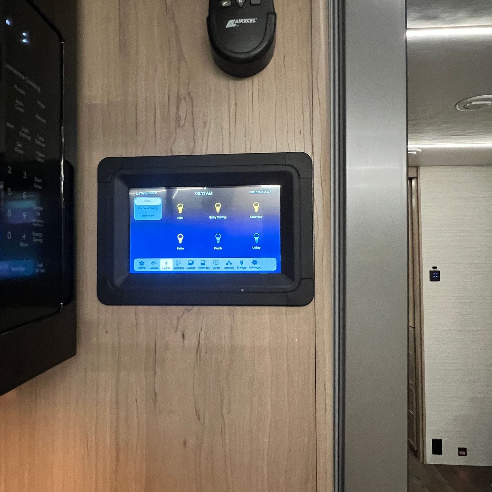 2027 Dynamax Europa S 37CA wall-mounted touchscreen control panel showing the lighting page with cab, entry ceiling, courtesy, patio, porch and utility zones, beside the microwave keypad and a thermostat remote