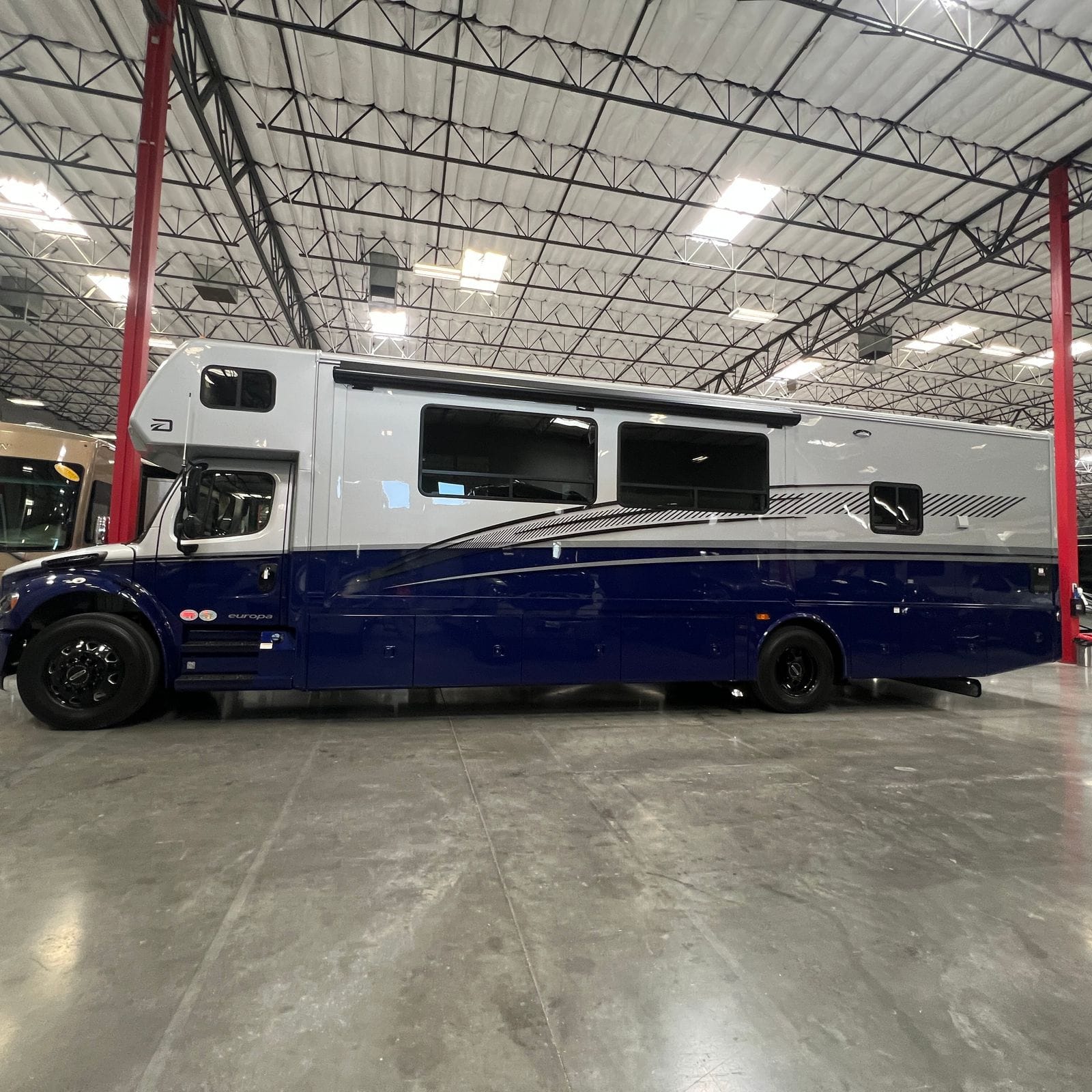 2027 Dynamax Europa S 37CA in Cobalt blue, full driver-side profile with blue lower half and silver upper, black swoosh graphics, awning rail over the slide, entry steps and a row of pass-through storage bay doors
