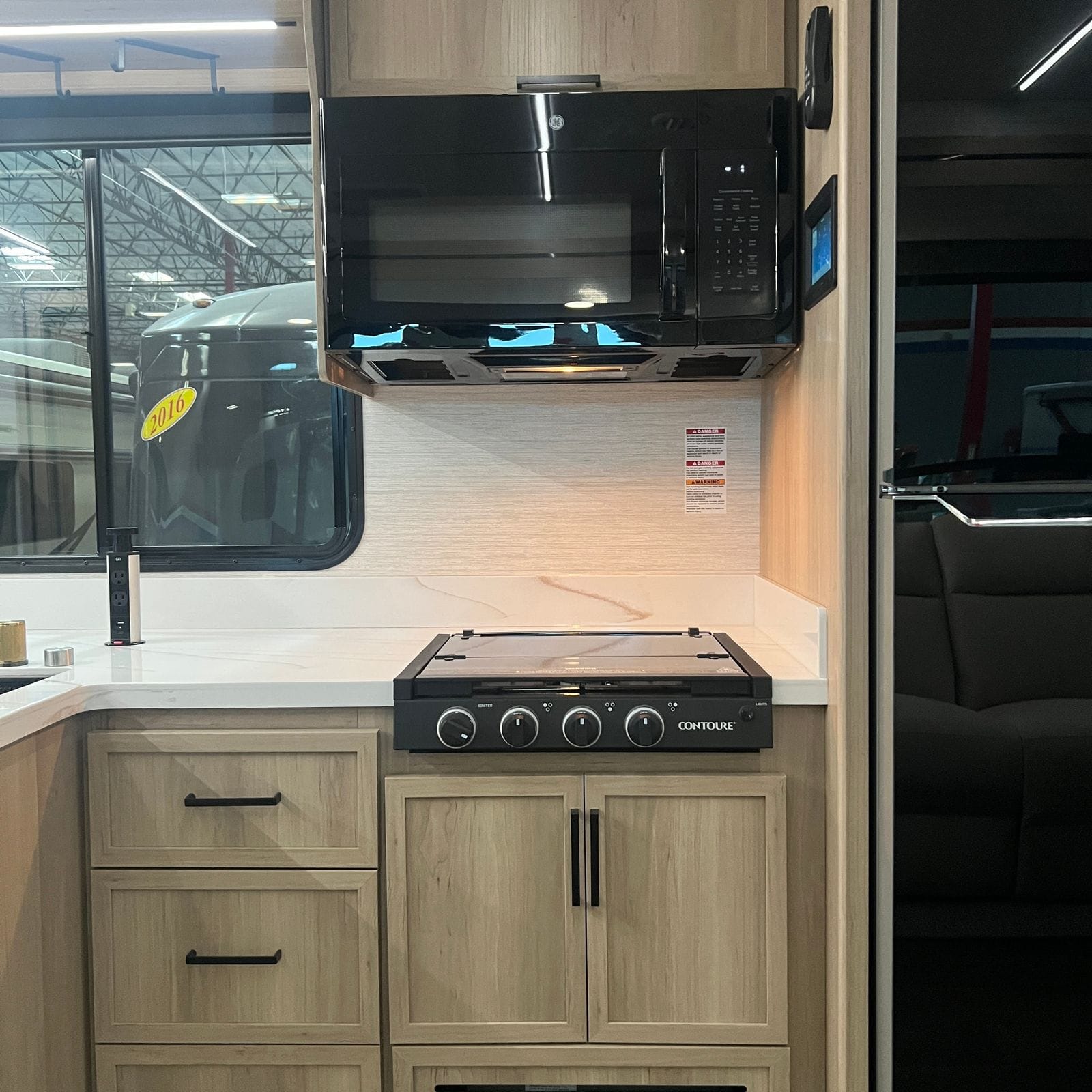 2027 Dynamax Europa S 37CA galley with an over-range microwave, three-burner cooktop with glass cover, white marbled countertop and backsplash and light wood shaker cabinets with black bar pulls