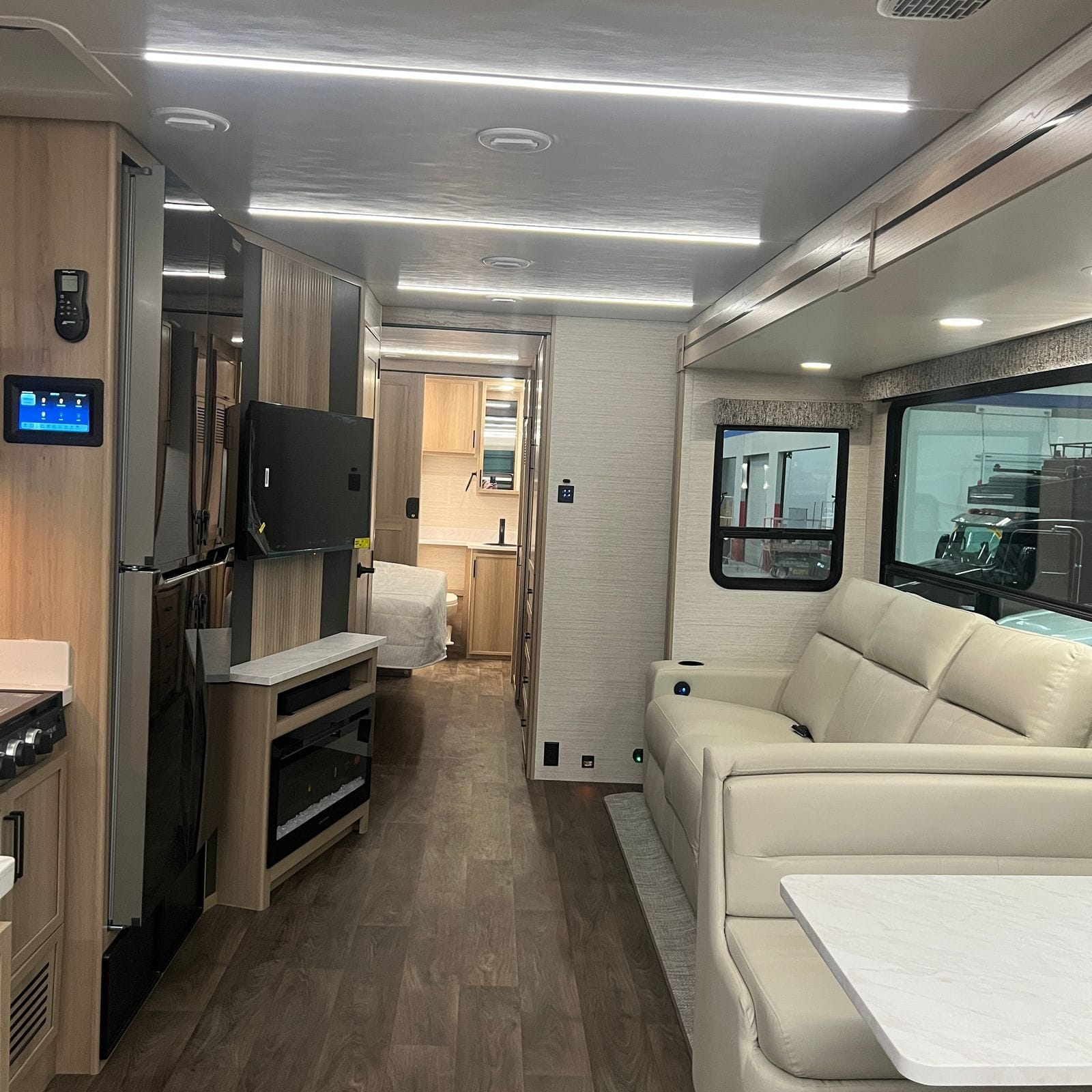2027 Dynamax Europa S 37CA interior looking rearward down the aisle with Alabaster leather sofa, wall-mounted television over an electric fireplace console, light wood cabinetry and wood-look vinyl flooring