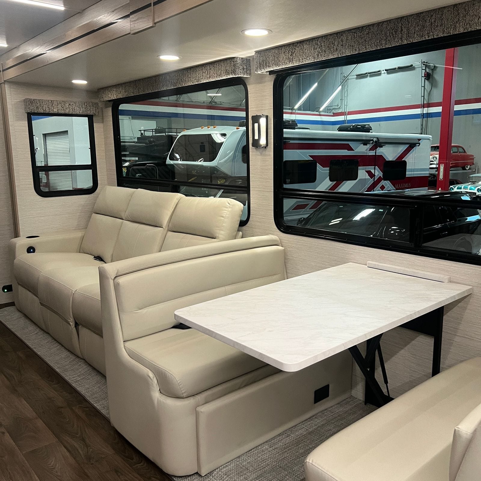 2027 Dynamax Europa S 37CA slide-out living area with an Alabaster leather power sofa, a marble-look free-standing table on a black pedestal base and large tinted windows with fabric valances