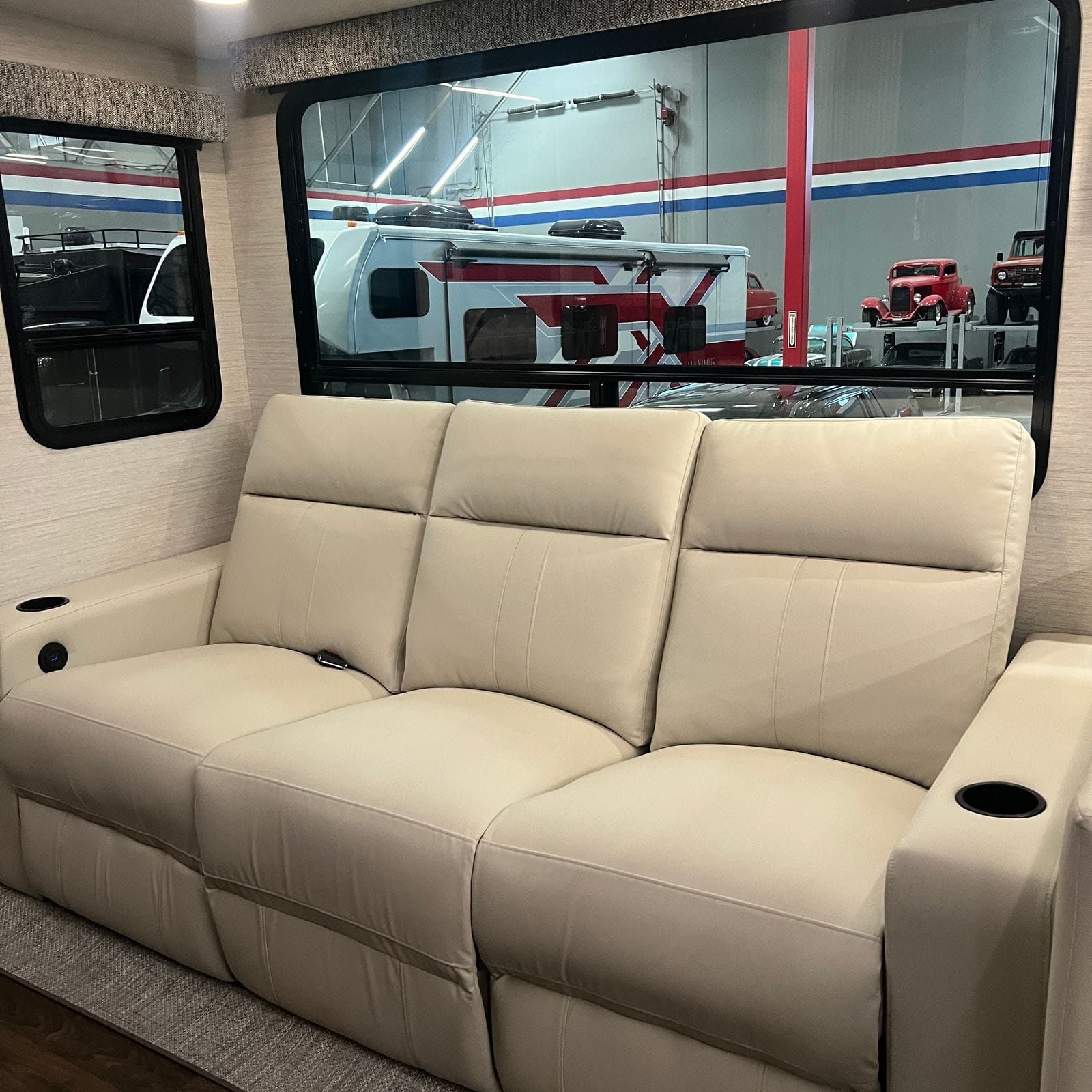 2027 Dynamax Europa S 37CA Alabaster leather three-seat power reclining sofa with cup-holder armrests below a large tinted slide-out window