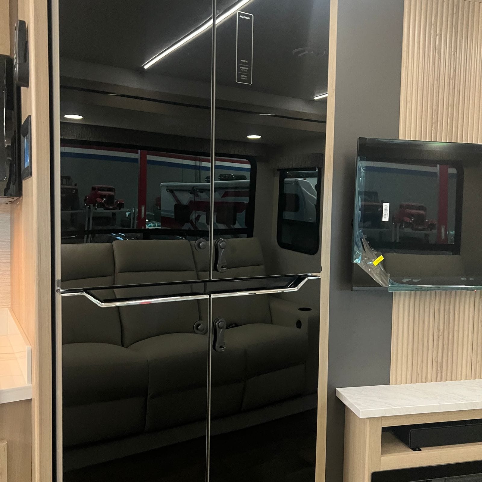 2027 Dynamax Europa S 37CA tall black french-door residential-style refrigerator flanked by light wood cabinet columns, with a flat-screen television on the fluted wood wall alongside