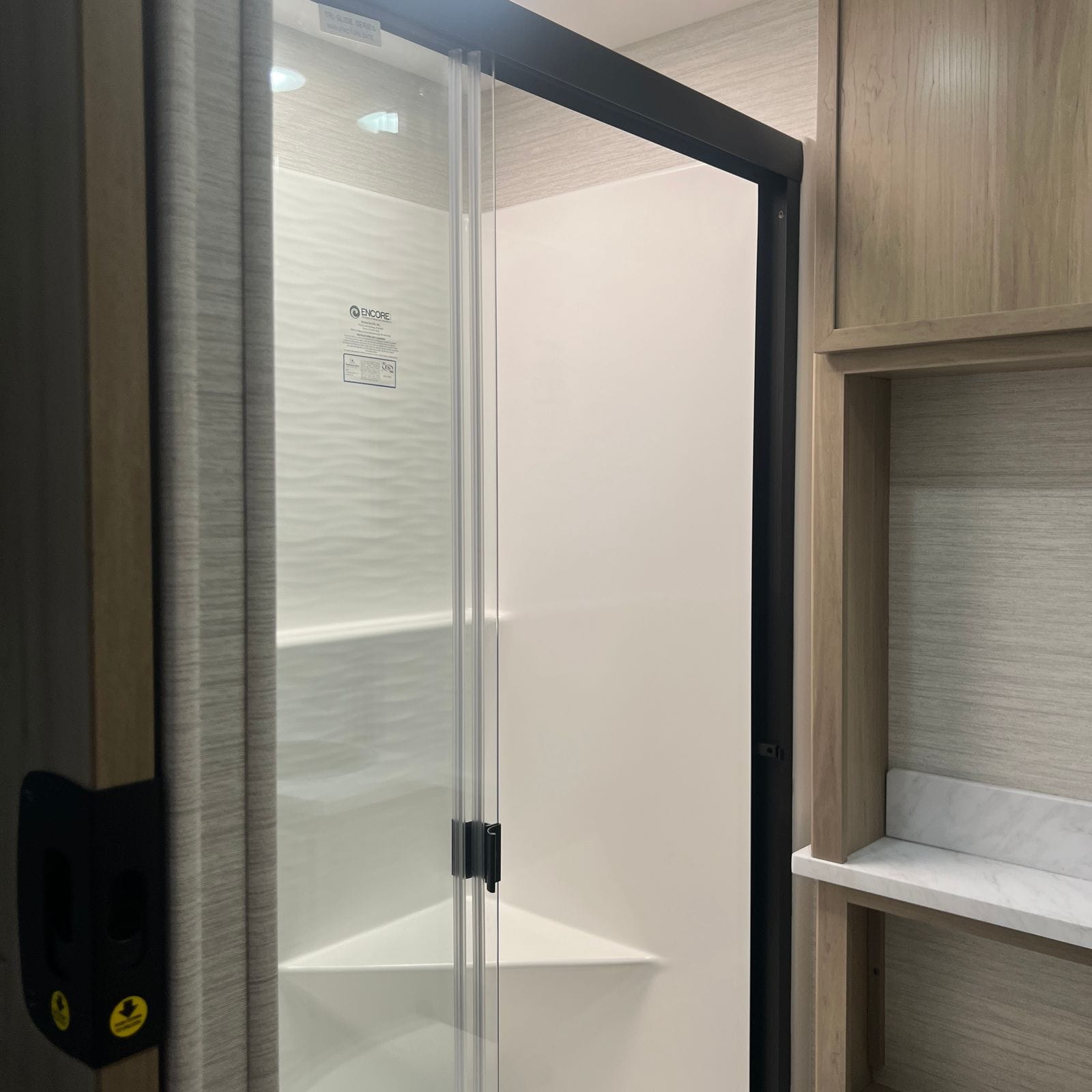 2027 Dynamax Europa S 37CA shower enclosure with black-framed clear glass doors, a white textured wave-pattern surround and a light wood shelf niche