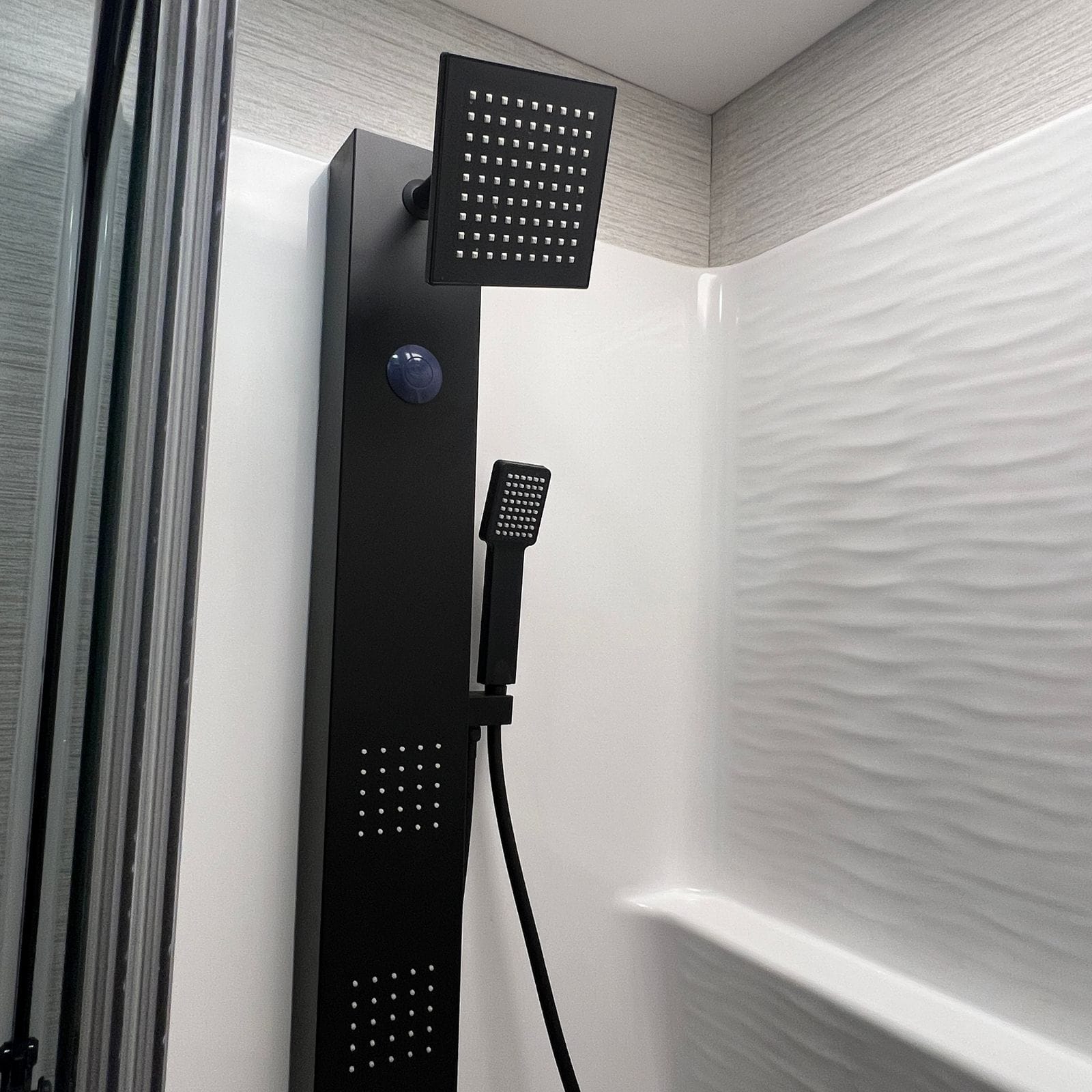 2027 Dynamax Europa S 37CA shower interior detail showing a matte black shower tower with a square rain head, handheld wand and body-spray jets against the wave-textured surround
