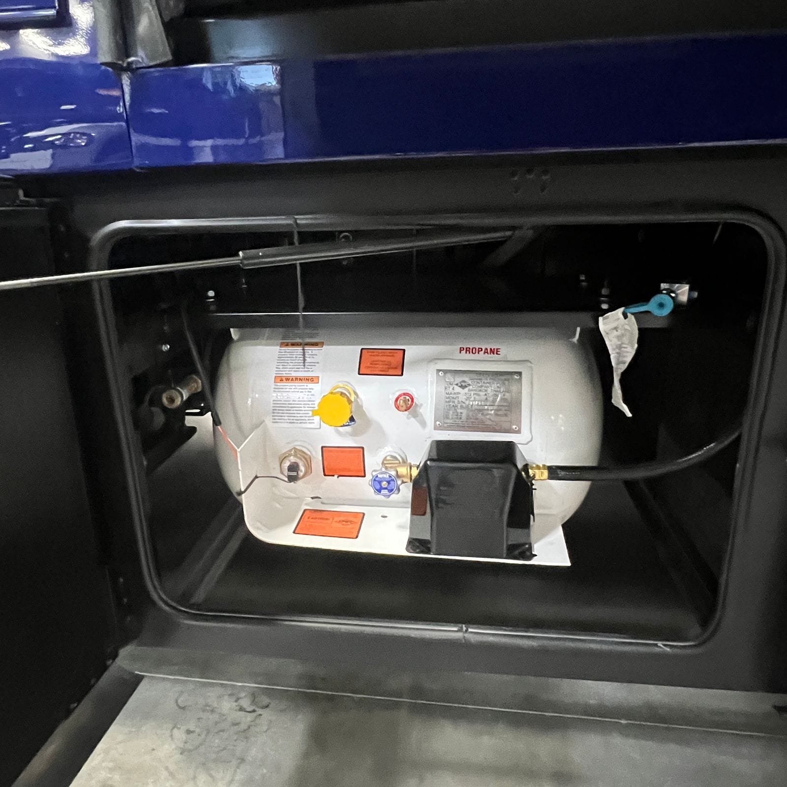 2027 Dynamax Europa S 37CA open propane compartment with a horizontal propane tank, regulator and brass fittings