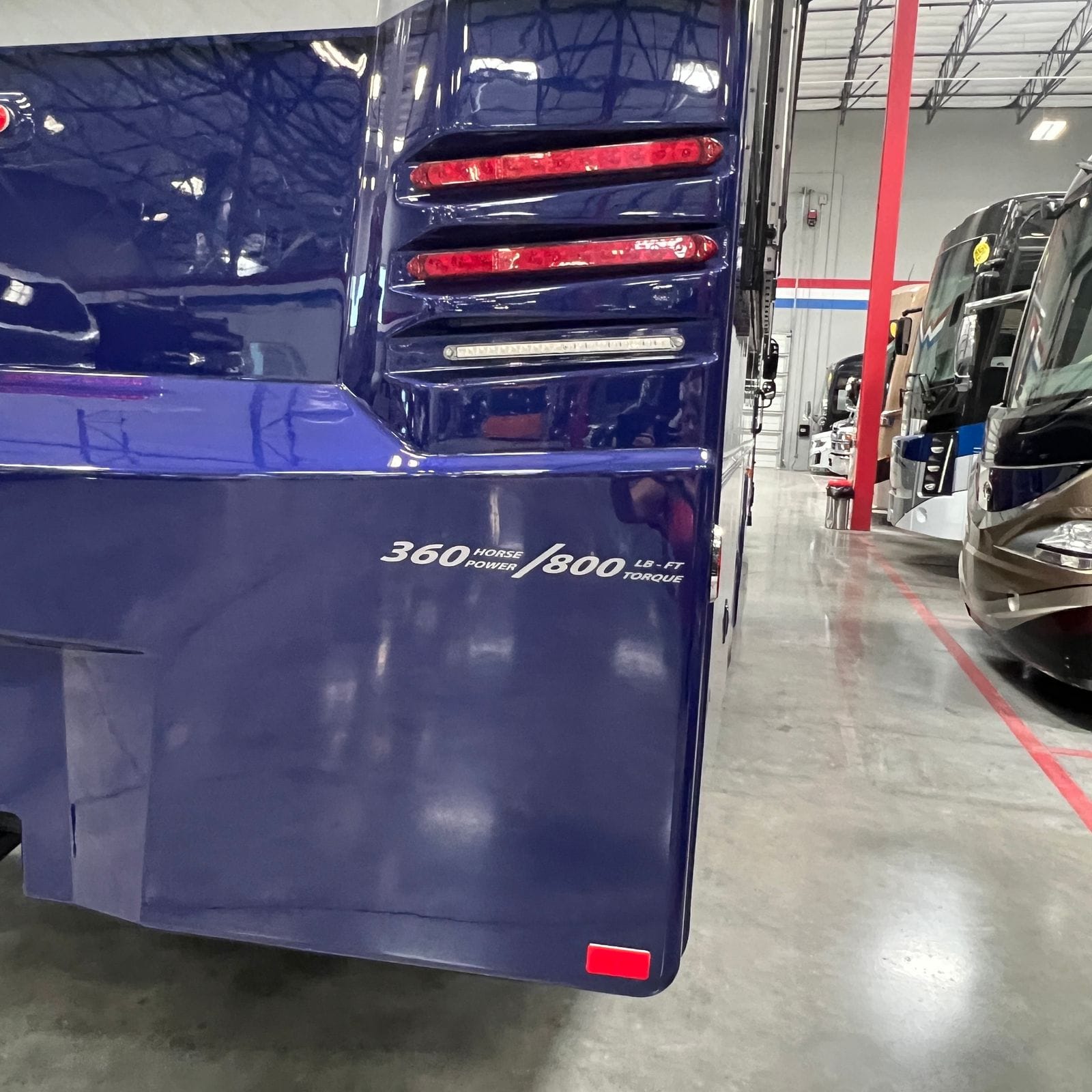 2027 Dynamax Europa S 37CA rear passenger-side corner detail showing the glossy blue panel, stacked red LED tail lamps, white reverse light strip and the 360 horsepower 800 lb-ft torque decal