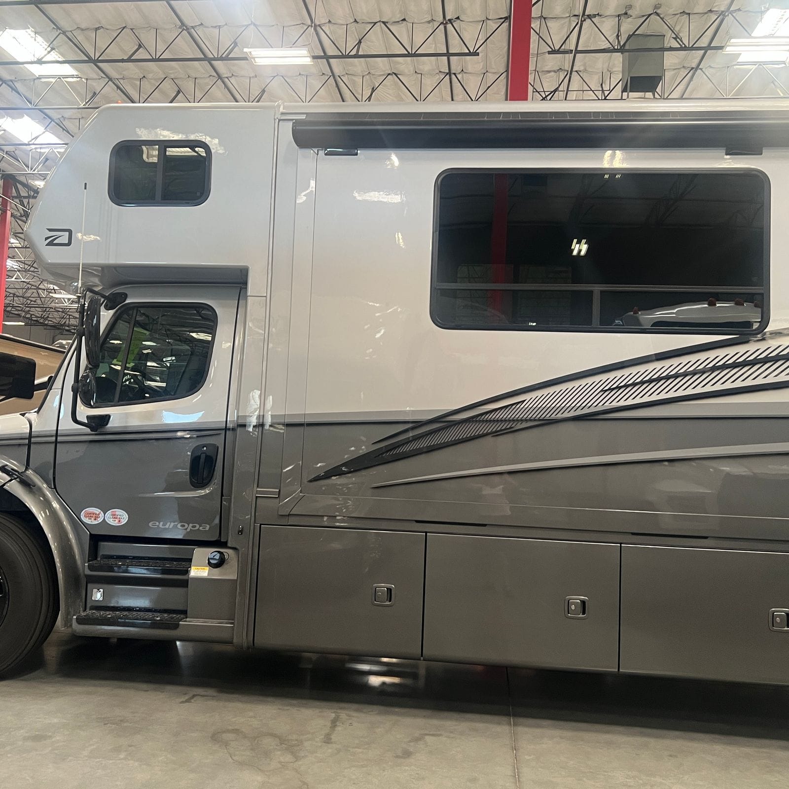 2027 Dynamax Europa S 37CA in Quicksilver, full driver-side profile showing the white upper cab-over, grey lower body with black and silver swoosh graphics, slide-out window and three pass-through storage bay doors