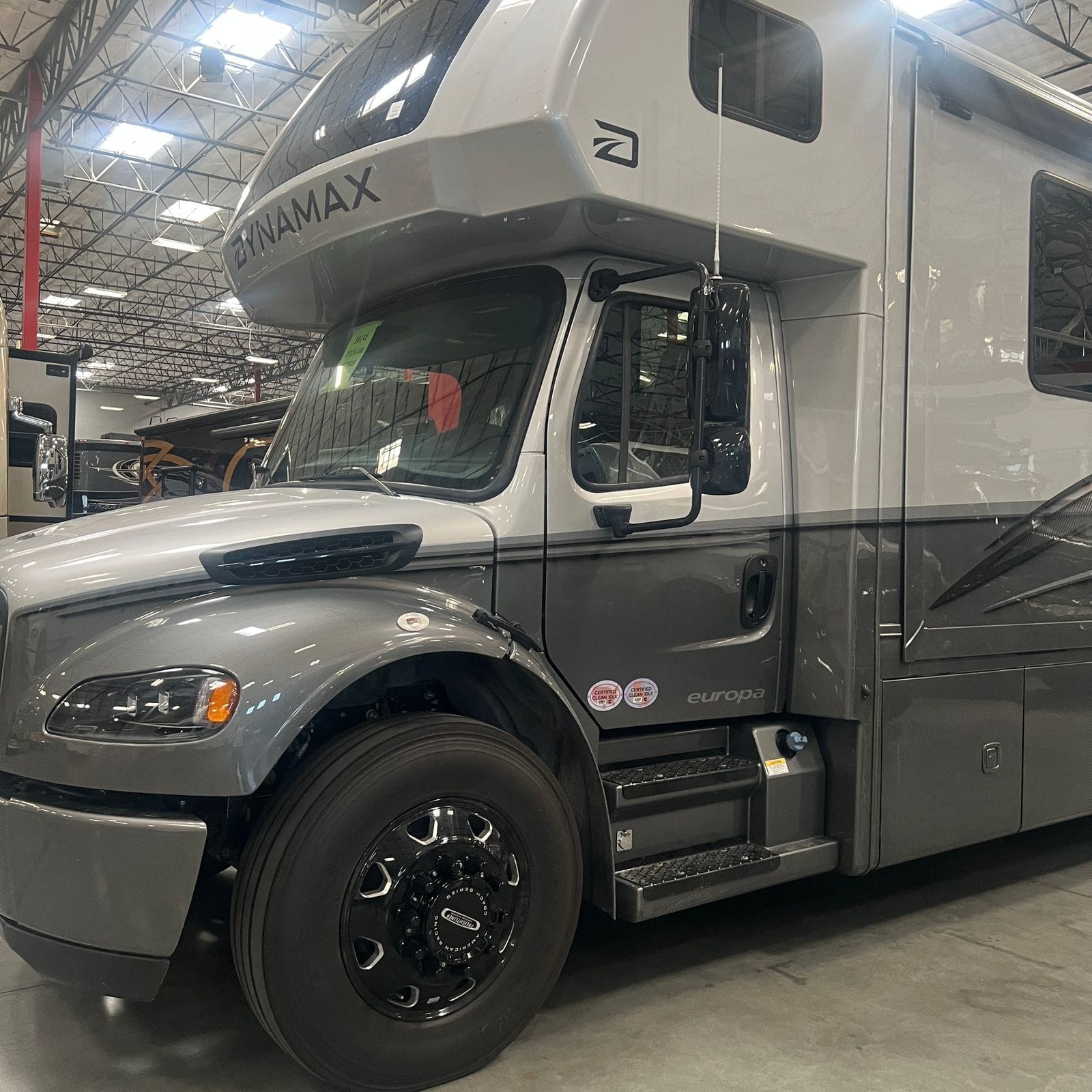 2027 Dynamax Europa S 37CA in Quicksilver, front driver-side three-quarter view showing the silver and charcoal grey Freightliner cab, black Dynamax badging, chrome-trimmed grille and black coach wheels
